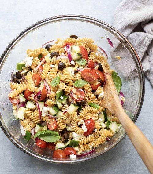 Pasta salad with vegetables in a glass bowl with a wooden spoon and grey cloth beside bowl. 